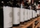A row of wine bottles is neatly arranged on a wooden shelf, with each bottle featuring a minimalist white label and a stylized logo. The focus is on the bottles in the foreground, while additional bottles blur into the background, suggesting a well-stocked collection.