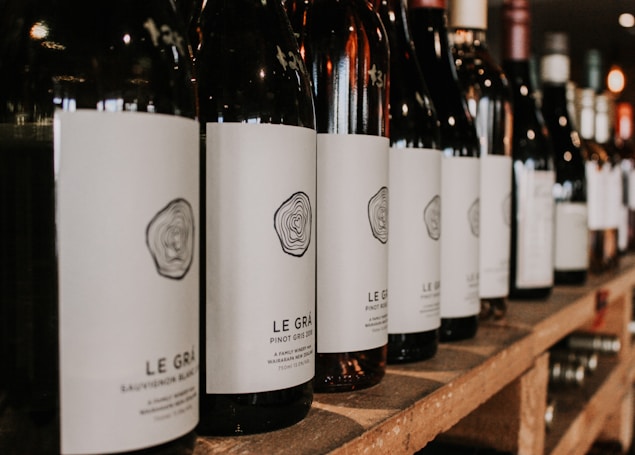 A row of wine bottles is neatly arranged on a wooden shelf, with each bottle featuring a minimalist white label and a stylized logo. The focus is on the bottles in the foreground, while additional bottles blur into the background, suggesting a well-stocked collection.
