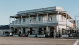 A two-story historic hotel building with a veranda and classic architectural design, featuring white wooden paneling and balconies. There are outdoor seating areas with tables and chairs under umbrellas, surrounded by potted plants.