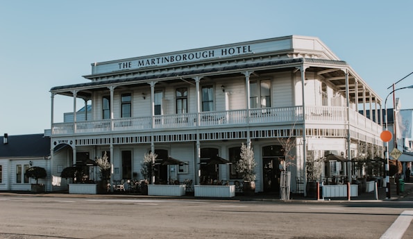 A two-story historic hotel building with a veranda and classic architectural design, featuring white wooden paneling and balconies. There are outdoor seating areas with tables and chairs under umbrellas, surrounded by potted plants.
