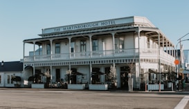 A two-story historic hotel building with a veranda and classic architectural design, featuring white wooden paneling and balconies. There are outdoor seating areas with tables and chairs under umbrellas, surrounded by potted plants.