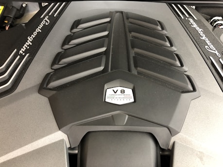 A close-up view of a Lamborghini engine cover, prominently featuring the V8 label and turbo specifications. The design includes a series of vents and angular lines, emphasizing a sleek and powerful appearance.