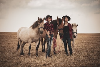 Two people dressed in casual western attire, including cowboy hats and plaid shirts, stand in a dry, open field with three horses. The scene depicts a rustic, rural atmosphere with overcast skies adding a muted tone.