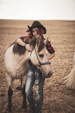 A person with red hair, wearing a plaid shirt and cowboy hat, stands in an open field with a light brown horse. The person affectionately leans on the horse, displaying a relaxed and friendly connection. The ground is covered in straw-colored stubble, suggesting a rural or farm setting.
