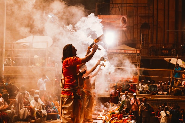 A group of people are performing a traditional ritual by the riverbank, surrounded by smoke and offerings. Attendees are seated in the background, observing the ceremony.