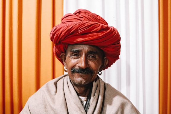 A man with a weathered face and a thick mustache is wearing a large, bright red turban and simple, traditional clothing. He is adorned with hoop earrings and poses against a background with vertical stripes in orange and white.