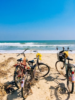 Several bicycles are parked on a sunny sandy beach with the ocean waves in the background. Three people are visible in the water, enjoying the waves. The sky is clear and blue, enhancing the bright and vibrant atmosphere.