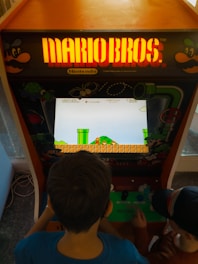 Two children are playing an arcade machine with a 'Mario Bros.' theme. The arcade cabinet features bright, colorful graphics typical of vintage video games, with the game screen displaying a classic Mario scene with green pipes and a pixelated landscape.