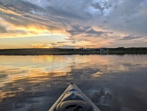 A serene lake view during sunset with golden and orange hues reflecting off the water's surface. The sky is adorned with dramatic clouds, and a small town with houses and trees can be seen on the horizon. A kayak is partially visible in the foreground, indicating a peaceful kayaking experience.