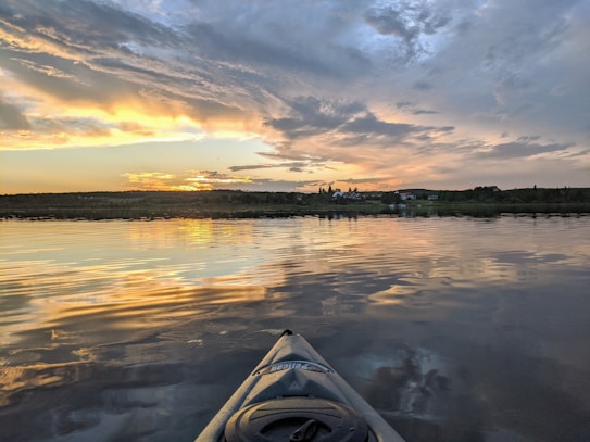 A serene lake view during sunset with golden and orange hues reflecting off the water's surface. The sky is adorned with dramatic clouds, and a small town with houses and trees can be seen on the horizon. A kayak is partially visible in the foreground, indicating a peaceful kayaking experience.
