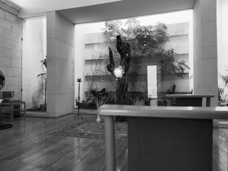 A spacious room featuring wooden flooring with furniture arranged around. In the center, a tree-like sculpture stands near a glass wall, behind which plants are visible, adding a touch of nature. The room appears well-lit with natural light coming through large windows.