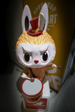 A whimsical character resembling a rabbit, with large ears and a fluffy body. It has wide eyes and a determined expression, wearing a small red top hat with a feather. The character is playing a colorful drum with two drumsticks.