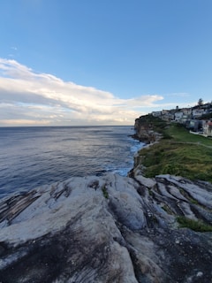 A coastal landscape featuring rocky cliffs leading down to the ocean. The clear blue sky meets the expansive sea on the horizon, with waves gently crashing against the rocks. A row of houses is visible along the cliff, basking in the natural light.