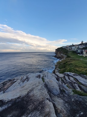 A coastal landscape featuring rocky cliffs leading down to the ocean. The clear blue sky meets the expansive sea on the horizon, with waves gently crashing against the rocks. A row of houses is visible along the cliff, basking in the natural light.