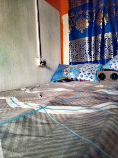 A bed covered with a patterned blanket in a corner of a room. The left wall is a muted grey, while the wall behind the bed features a vibrant mix of colors with blue and gold ornate curtains. A colorful pillow rests against the wall. There is an electrical socket with a plugged-in charger on the wall next to the bed.