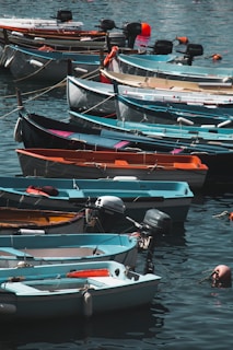 A collection of small boats neatly lined up along a calm waterfront. Each boat varies in color, including shades of blue, white, orange, and teal. They are equipped with outboard motors and some have buoyant life preservers attached. The water reflects the colorful hues of the boats and gently ripples around them.