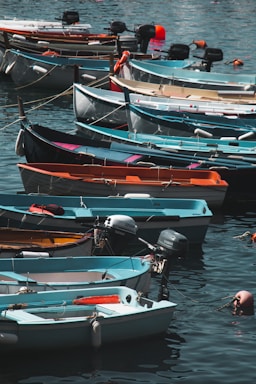 A collection of small boats neatly lined up along a calm waterfront. Each boat varies in color, including shades of blue, white, orange, and teal. They are equipped with outboard motors and some have buoyant life preservers attached. The water reflects the colorful hues of the boats and gently ripples around them.