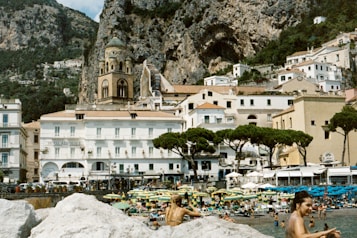 A picturesque coastal town with historic architecture nestled against rugged cliffs. The town features a prominent cathedral with a distinctive dome, surrounded by colorful buildings and lush greenery. In the foreground, people are enjoying a crowded beach with umbrellas and beach chairs.