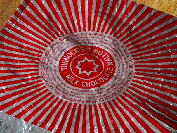 A crumpled aluminum wrapper featuring a striking red and silver design with radial stripes extending from a central circle. The circle contains the text 'Tunnock's Milk Chocolate', along with additional smaller text detailing ingredients and manufacturer information. The geometric pattern draws attention with its bold and symmetrical arrangement.