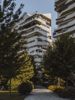 A modern residential building with a unique architectural design, featuring multiple balconies and terraces with greenery. The structure is predominantly white with dark accents. It is surrounded by lush green trees, creating a serene and natural environment. A paved pathway leads to the building, enhancing the garden-like setting.