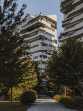 A modern residential building with a unique architectural design, featuring multiple balconies and terraces with greenery. The structure is predominantly white with dark accents. It is surrounded by lush green trees, creating a serene and natural environment. A paved pathway leads to the building, enhancing the garden-like setting.