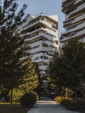 A modern residential building with a unique architectural design, featuring multiple balconies and terraces with greenery. The structure is predominantly white with dark accents. It is surrounded by lush green trees, creating a serene and natural environment. A paved pathway leads to the building, enhancing the garden-like setting.