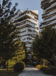 A modern residential building with a unique architectural design, featuring multiple balconies and terraces with greenery. The structure is predominantly white with dark accents. It is surrounded by lush green trees, creating a serene and natural environment. A paved pathway leads to the building, enhancing the garden-like setting.