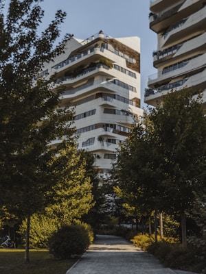 A modern residential building with a unique architectural design, featuring multiple balconies and terraces with greenery. The structure is predominantly white with dark accents. It is surrounded by lush green trees, creating a serene and natural environment. A paved pathway leads to the building, enhancing the garden-like setting.