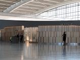 A modern airport interior with a high ceiling and large windows allowing natural light to illuminate the space. The area features a lounge or check-in area with a sleek, wooden and glass partition. Two suited individuals stand near a podium, while another person is standing to the right, possibly waiting or on the phone. The environment appears calm and professional.