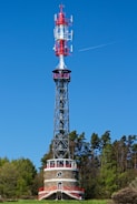 A tall communication tower with various antennas and devices is situated in a green forested area. The base of the tower appears to be made of stone and features red accents and a small window. The sky is clear and blue, with a visible contrail from an aircraft.