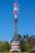 A tall communication tower with various antennas and devices is situated in a green forested area. The base of the tower appears to be made of stone and features red accents and a small window. The sky is clear and blue, with a visible contrail from an aircraft.