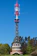 A tall communication tower with various antennas and devices is situated in a green forested area. The base of the tower appears to be made of stone and features red accents and a small window. The sky is clear and blue, with a visible contrail from an aircraft.