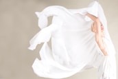 A person is gracefully dancing with a large, flowing piece of white fabric, creating a sense of movement and elegance. The background is a soft, neutral color that enhances the contrast and fluidity of the fabric.