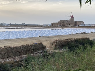 A windmill with a red cap is situated near a building by a large expanse of land divided into rectangular sections. The area is covered with heaps of white salt, indicating a salt farm. In the foreground, there is a stack of wooden planks and fencing, with vegetation growing around it.