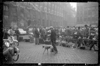 A bustling street market scene from the past, with vendors and buyers interacting amidst a range of goods displayed on tables. People are dressed in vintage attire, and a dog walks in the foreground. In the background, historical buildings line the street, and classic cars are parked along the side.