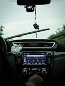 The interior view of a car showing a dashboard with a digital display tuned to a radio station and various controls. A hand is visible near the temperature controls, and there is a hanging ornament attached to the rearview mirror. The windshield wipers are slightly elevated, indicating recent use.