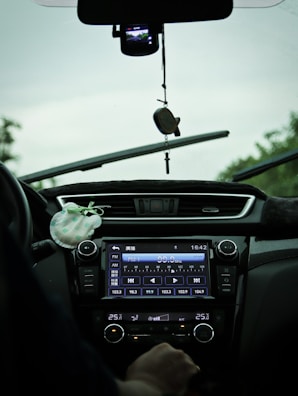 The interior view of a car showing a dashboard with a digital display tuned to a radio station and various controls. A hand is visible near the temperature controls, and there is a hanging ornament attached to the rearview mirror. The windshield wipers are slightly elevated, indicating recent use.