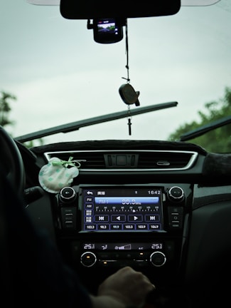 The interior view of a car showing a dashboard with a digital display tuned to a radio station and various controls. A hand is visible near the temperature controls, and there is a hanging ornament attached to the rearview mirror. The windshield wipers are slightly elevated, indicating recent use.