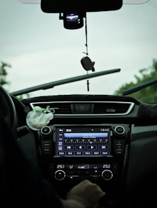 The interior view of a car showing a dashboard with a digital display tuned to a radio station and various controls. A hand is visible near the temperature controls, and there is a hanging ornament attached to the rearview mirror. The windshield wipers are slightly elevated, indicating recent use.