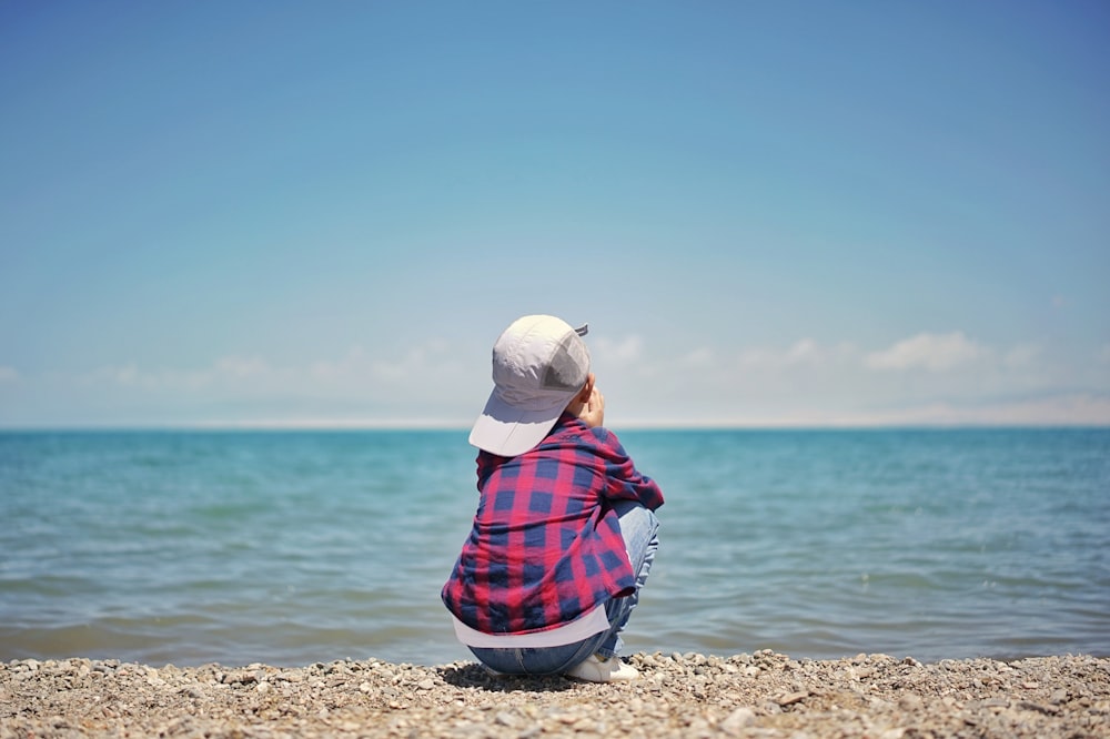 낮 동안 수역 근처의 갈색 모래 위에 앉아 있는 흰색 니트 모자를 쓴 여자