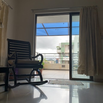 A room with an open sliding glass door leading to a balcony covered by a blue canopy. Inside the room, there is a dark wooden rocking chair with a cushion and a window with cream-colored curtains. Outside, there's a railing and a view of modern apartment buildings with balconies.
