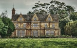 An elegant large manor house with distinct Gothic architectural features stands surrounded by lush, dense trees and greenery. The structure has a triangular gabled roof with multiple windows on each of its three floors, and a clock tower is visible on the left side of the building. The facade shows an intricate design with a combination of stone and traditional elements.