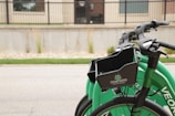 Three green bicycles with baskets are lined up on a street. The bicycles have the logo 'CEDAR RAPIDS City of Five Seasons' on the baskets. A concrete sidewalk and building with windows and a railing are visible in the background.