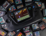 A Sega Game Gear handheld console prominently displays a game on its screen, surrounded by a variety of Game Gear cartridges featuring colorful artwork. The game in play depicts a blue character, possibly Sonic, navigating a platform environment. The console has a black design with control buttons and a directional pad.