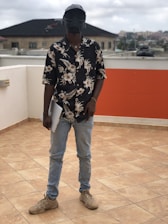 A person stands on a tiled rooftop holding a laptop. They are wearing a floral-patterned shirt, light blue jeans, and beige sneakers. The individual has a unique helmet or mask that resembles a futuristic or sci-fi design. The background features residential buildings and a cloudy sky.