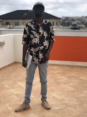 A person stands on a tiled rooftop holding a laptop. They are wearing a floral-patterned shirt, light blue jeans, and beige sneakers. The individual has a unique helmet or mask that resembles a futuristic or sci-fi design. The background features residential buildings and a cloudy sky.