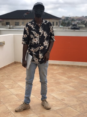 A person stands on a tiled rooftop holding a laptop. They are wearing a floral-patterned shirt, light blue jeans, and beige sneakers. The individual has a unique helmet or mask that resembles a futuristic or sci-fi design. The background features residential buildings and a cloudy sky.