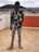 A person stands on a tiled rooftop holding a laptop. They are wearing a floral-patterned shirt, light blue jeans, and beige sneakers. The individual has a unique helmet or mask that resembles a futuristic or sci-fi design. The background features residential buildings and a cloudy sky.