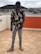 A person stands on a tiled rooftop holding a laptop. They are wearing a floral-patterned shirt, light blue jeans, and beige sneakers. The individual has a unique helmet or mask that resembles a futuristic or sci-fi design. The background features residential buildings and a cloudy sky.