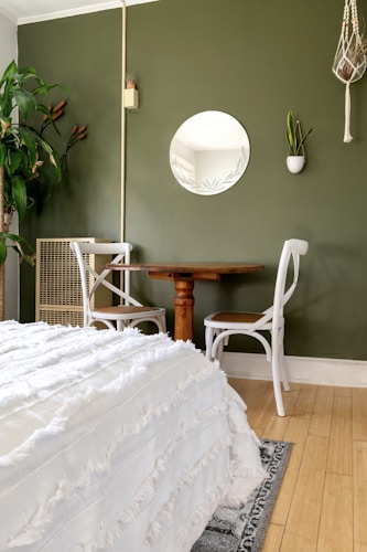 A cozy, minimalist room features a green wall adorned with a circular mirror and a small potted plant. A wood table is accompanied by two white chairs, and a large leafy plant adds a touch of nature. A white, textured bedspread extends over the bed, contrasting with the warm wood floor.